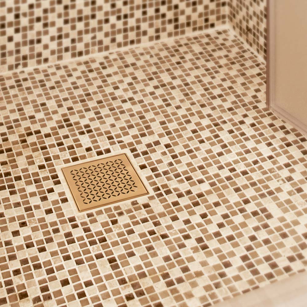 QM Drain Delmar Lotus Square Drain in Gold finish, installed in a luxury bathroom with a floral-pattern grate design. The image showcases the elegant stainless-steel grate, seamless tile integration, and efficient water drainage, offering a sophisticated and modern aesthetic for high-end shower and bathroom installations.
