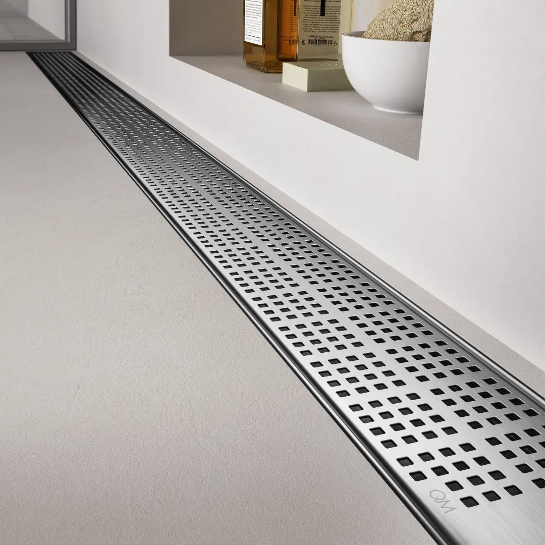 Modern spa-style bathroom scene featuring the QM Drain Kai Linear Drain—sleek stainless-steel channel set into large-format porcelain tiles, with integrated waterproof flange beneath the tile edges, minimalist grate design, and rapid high-flow drainage for a seamless luxury shower aesthetic.